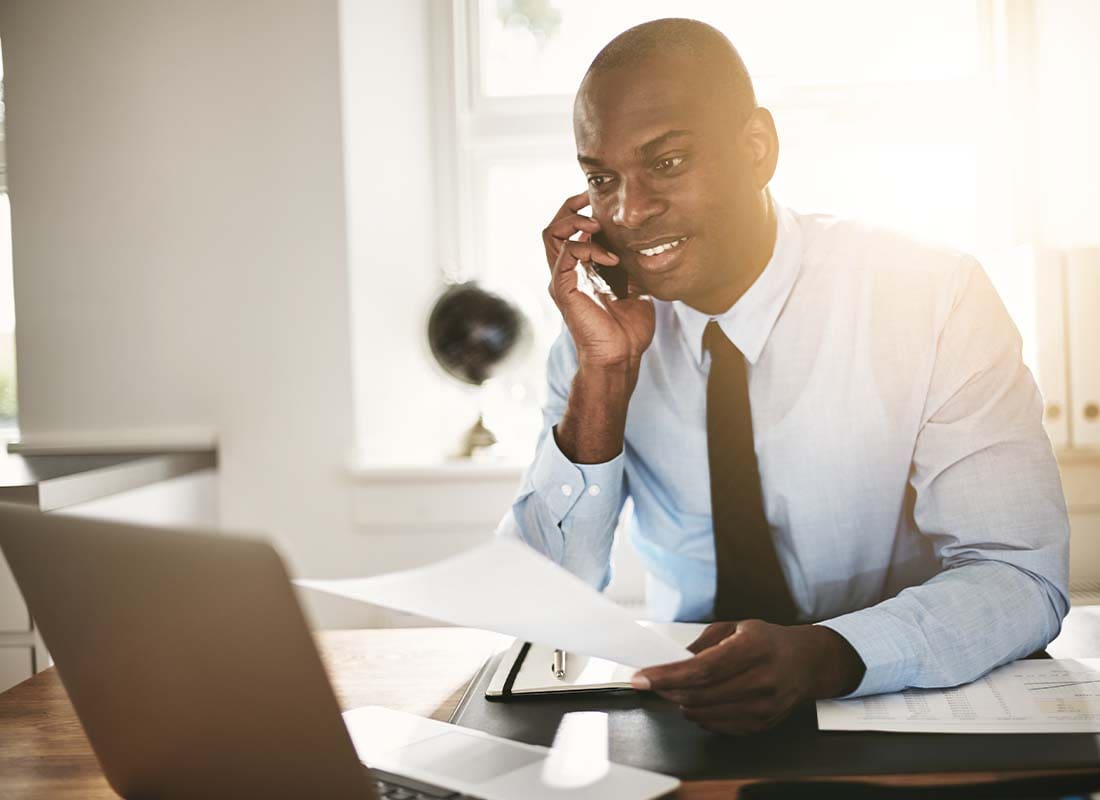 Surety Bond - Smiling Businessman Examining Documentation over the Phone and Working from His Home Office and Using a Laptop