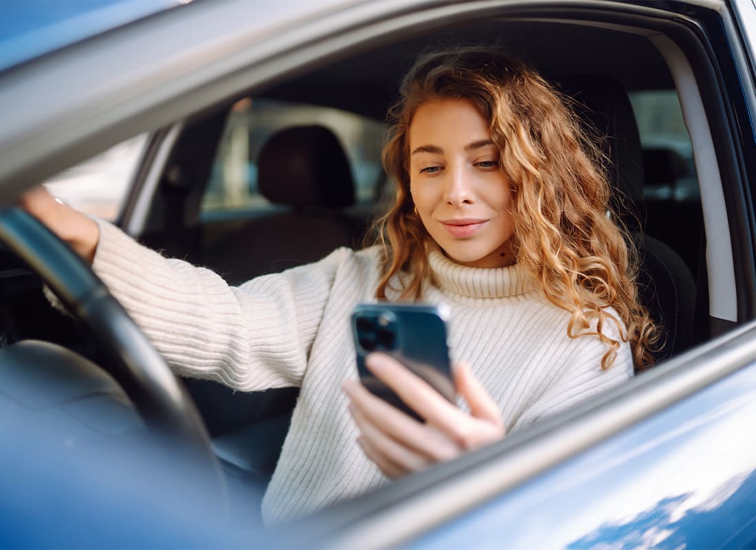 Report a Claim - Young Woman Sitting in a Car in the Driver’s Seat Looking at a Smartphone